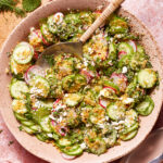A vibrant salad of sliced cucumbers, radishes, and fresh chopped herbs is garnished with crumbled feta cheese and a sprinkle of breadcrumbs. The salad is presented in a rustic pink bowl with a serving spoon, set on a soft pink textured surface.
