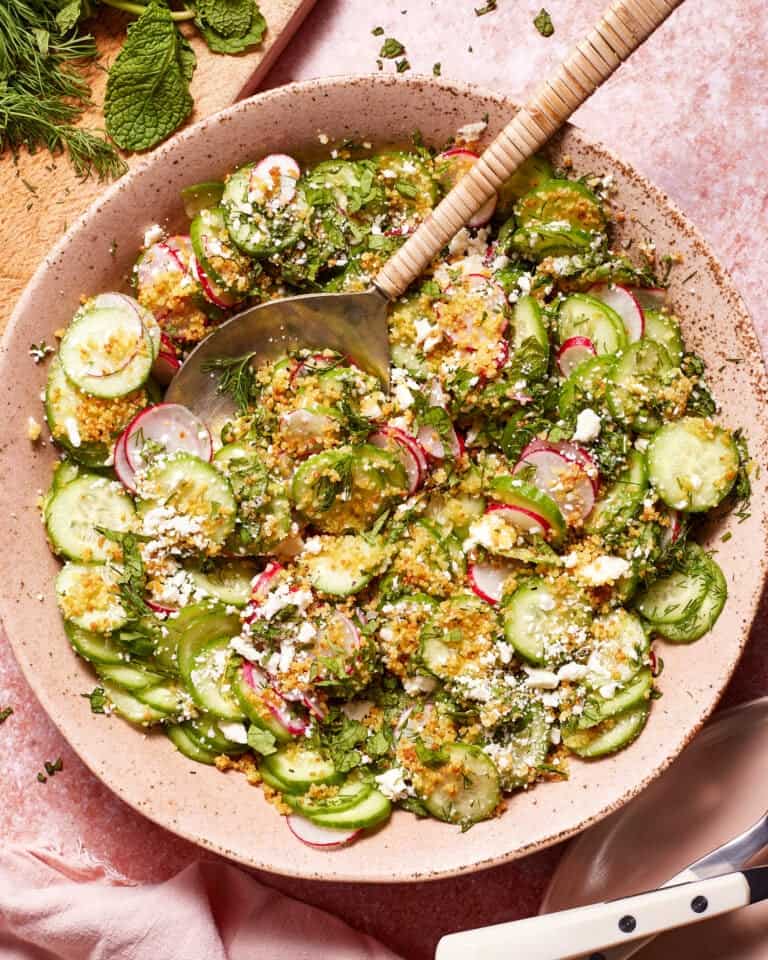 A vibrant salad of sliced cucumbers, radishes, and fresh chopped herbs is garnished with crumbled feta cheese and a sprinkle of breadcrumbs. The salad is presented in a rustic pink bowl with a serving spoon, set on a soft pink textured surface.