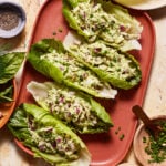 A plate of four lettuce wraps filled with a creamy chicken salad mixture, garnished with chopped chives. Surrounding the plate are small bowls containing ground pepper and chopped herbs. The background features a marble surface with a variety of green leaves.
