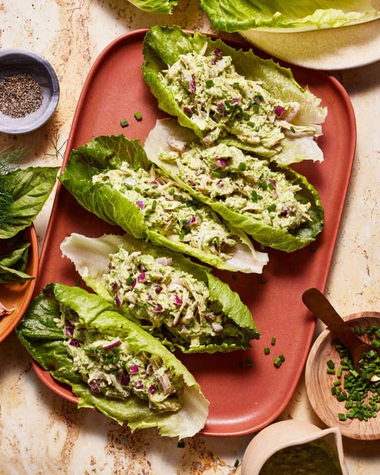 A plate of four lettuce wraps filled with a creamy chicken salad mixture, garnished with chopped chives. Surrounding the plate are small bowls containing ground pepper and chopped herbs. The background features a marble surface with a variety of green leaves.