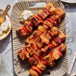 A platter holds several skewers of grilled meat and vegetables, featuring chunks of seasoned meat, red peppers, and red onions. In the background are a small dish of dip with seasoning, a bowl with a spoon, and part of a flatbread.