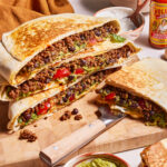 A close-up of a wooden board filled with cheesy taco melts stuffed with ground beef, black beans, tomatoes, lettuce, and melted cheese, served with bowls of guacamole and black beans, a knife, and a bottle of hot sauce in the background.