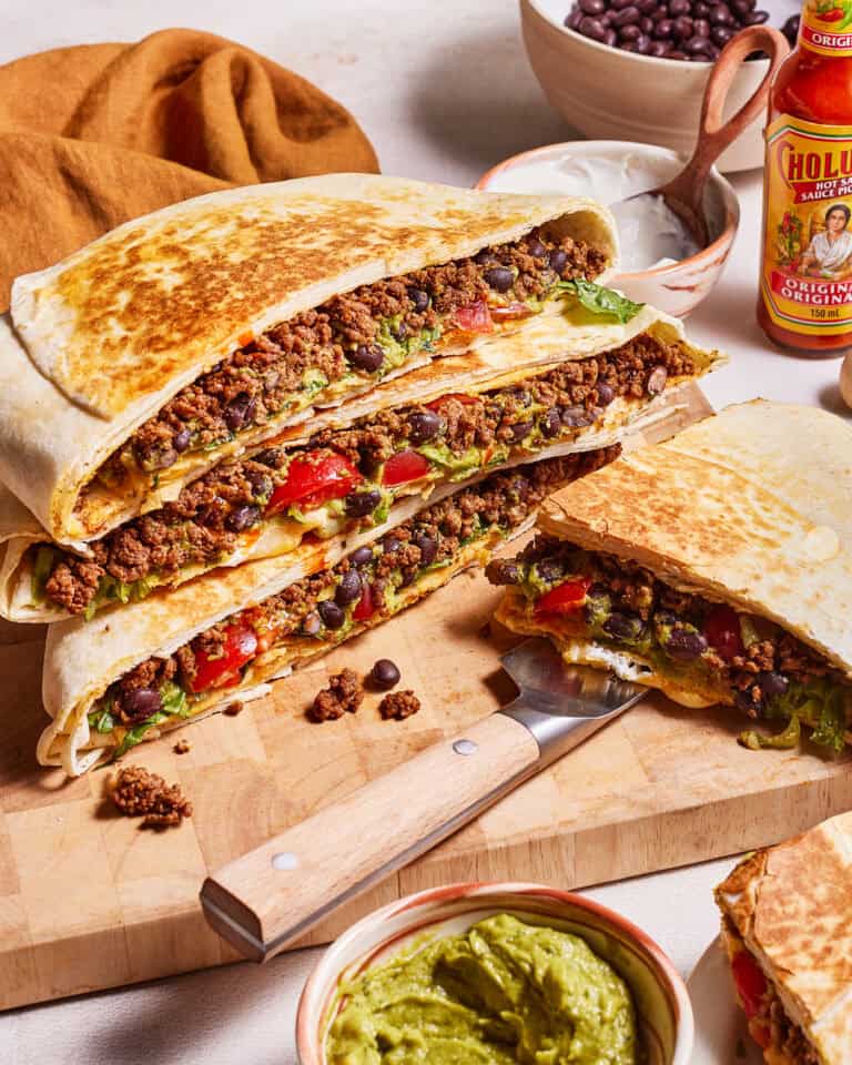 A close-up of a wooden board filled with cheesy taco melts stuffed with ground beef, black beans, tomatoes, lettuce, and melted cheese, served with bowls of guacamole and black beans, a knife, and a bottle of hot sauce in the background.