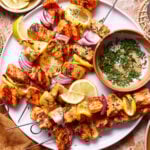A plate of grilled salmon skewers with red onions and lemon slices, garnished with chopped herbs. A small bowl filled with fresh herbs is placed on the plate. The meal is presented on a marble surface with lemon wedges on the side and a brown napkin.