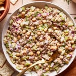 A large white bowl filled with a mixed salad containing chickpeas, celery, tuna, red onions, and herbs. There is a gold spoon in the bowl, and some crackers are visible in the background. The dish is set on a tiled surface.