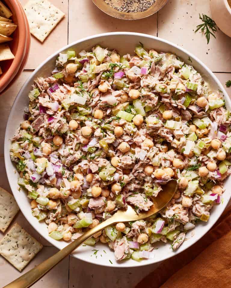 A large white bowl filled with a mixed salad containing chickpeas, celery, tuna, red onions, and herbs. There is a gold spoon in the bowl, and some crackers are visible in the background. The dish is set on a tiled surface.