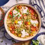 A large pot of pasta topped with cherry tomatoes, dollops of ricotta cheese, and fresh basil sits on a marble countertop. Surrounding the pot are a wooden cutting board with chopped basil, a bowl of grated cheese, a striped towel, and a glass of wine.