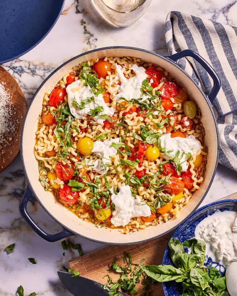 A large pot of pasta topped with cherry tomatoes, dollops of ricotta cheese, and fresh basil sits on a marble countertop. Surrounding the pot are a wooden cutting board with chopped basil, a bowl of grated cheese, a striped towel, and a glass of wine.