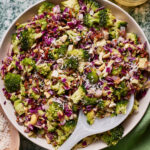 A bowl of colorful broccoli salad featuring chopped broccoli, red cabbage, nuts, sunflower seeds, and shredded cheese. A white serving spoon is placed in the bowl. The salad is arranged on a green marble surface with a small bowl of dressing nearby.