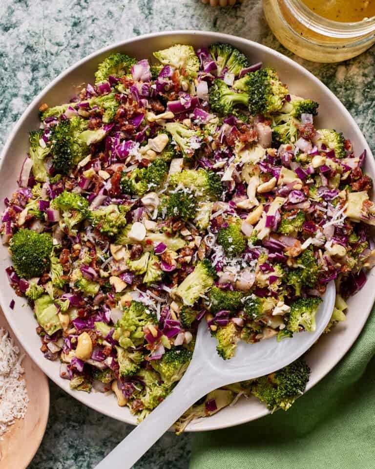 A bowl of colorful broccoli salad featuring chopped broccoli, red cabbage, nuts, sunflower seeds, and shredded cheese. A white serving spoon is placed in the bowl. The salad is arranged on a green marble surface with a small bowl of dressing nearby.