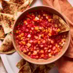 A bowl of colorful fruit salsa with diced strawberries, kiwi, and other fruits, surrounded by cinnamon-dusted tortilla chips on a white platter. An orange cloth is draped to the side.