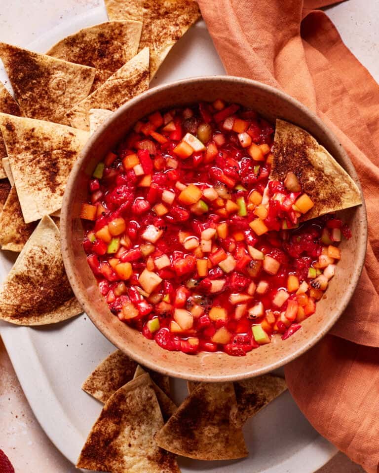 A bowl of colorful fruit salsa with diced strawberries, kiwi, and other fruits, surrounded by cinnamon-dusted tortilla chips on a white platter. An orange cloth is draped to the side.