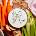 A platter with sliced carrot sticks, celery sticks, and round toasted bread crisps surrounding a bowl of creamy dip topped with chopped herbs. A small bowl with additional chopped herbs is in the background, beside a pink napkin.