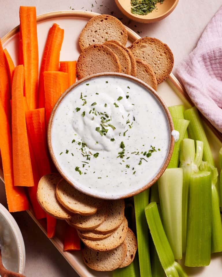 A platter with sliced carrot sticks, celery sticks, and round toasted bread crisps surrounding a bowl of creamy dip topped with chopped herbs. A small bowl with additional chopped herbs is in the background, beside a pink napkin.