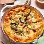 A savory vegetable quiche in a white baking dish, featuring golden, flaky pastry edges and a colorful filling of zucchini slices, red bell pepper chunks, and herbs. A slice has been cut out, revealing its hearty, cheesy interior.