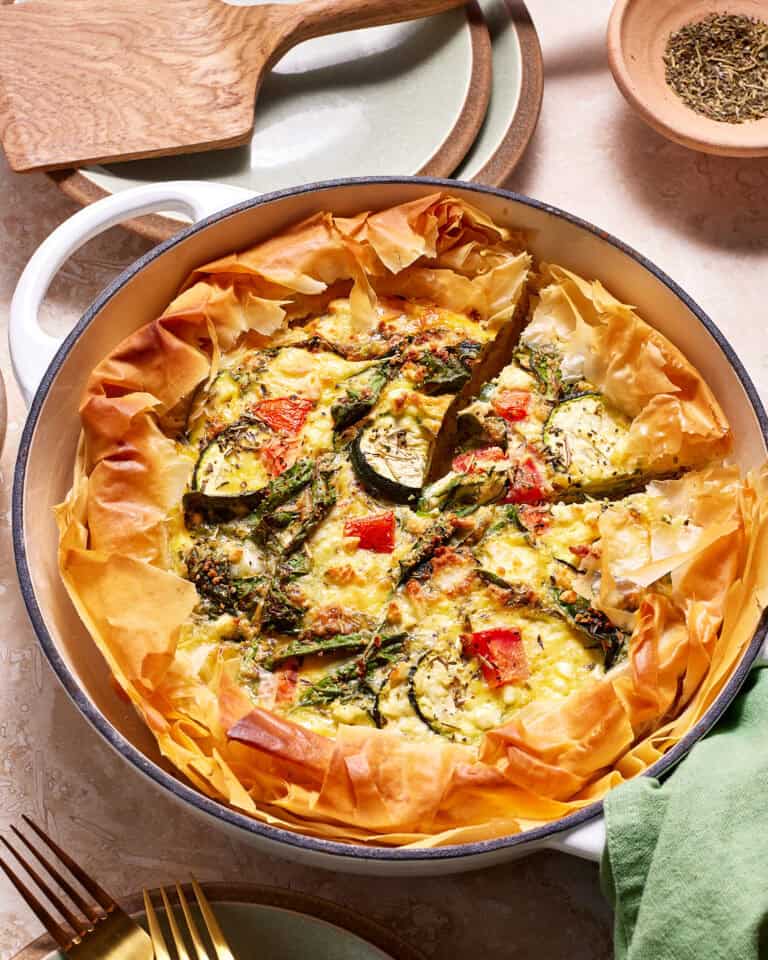 A savory vegetable quiche in a white baking dish, featuring golden, flaky pastry edges and a colorful filling of zucchini slices, red bell pepper chunks, and herbs. A slice has been cut out, revealing its hearty, cheesy interior.