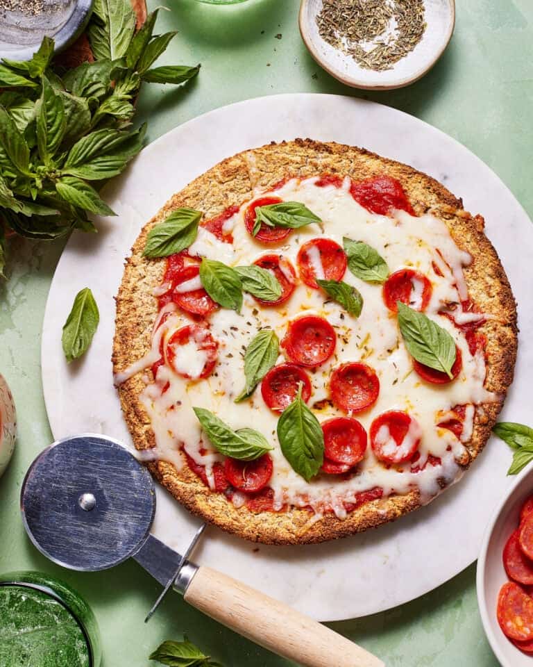 A cauliflower crust pizza topped with melted mozzarella cheese, pepperoni slices, and fresh basil leaves. The pizza is placed on a round marble board, with a pizza cutter nearby. Surrounding the pizza are bowls containing spices and fresh basil.