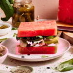 A creative sandwich made with thick slices of watermelon instead of bread, layered with greens, cheese, pickles, and meats, garnished with basil leaves, and drizzled with balsamic vinegar. The sandwich is held together with a toothpick.