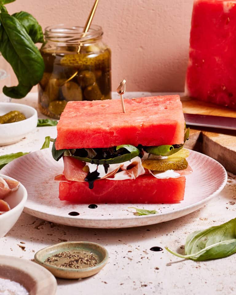 A creative sandwich made with thick slices of watermelon instead of bread, layered with greens, cheese, pickles, and meats, garnished with basil leaves, and drizzled with balsamic vinegar. The sandwich is held together with a toothpick.
