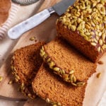 A loaf of pumpkin bread topped with pumpkin seeds is partially sliced on a wooden cutting board. A couple of slices are laid out on the board, revealing the moist texture of the bread. A knife with a white handle rests next to the loaf. There is also a striped cloth and a glass jar in the background.