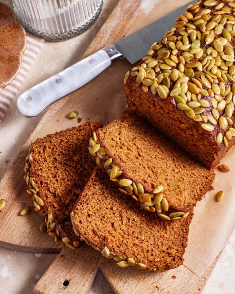 A loaf of pumpkin bread topped with pumpkin seeds is partially sliced on a wooden cutting board. A couple of slices are laid out on the board, revealing the moist texture of the bread. A knife with a white handle rests next to the loaf. There is also a striped cloth and a glass jar in the background.
