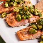A plate of grilled salmon fillets topped with a chunky avocado and herb salsa. The salmon has a slightly crispy, peppered exterior and the vibrant salsa adds a fresh, colorful garnish. A wooden spatula is partially visible on the right side of the plate.