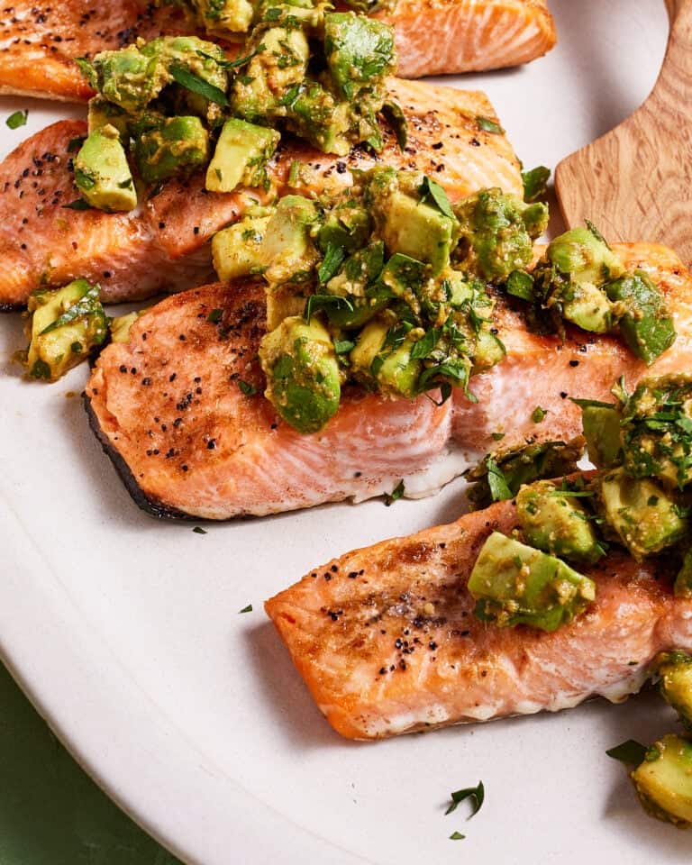 A plate of grilled salmon fillets topped with a chunky avocado and herb salsa. The salmon has a slightly crispy, peppered exterior and the vibrant salsa adds a fresh, colorful garnish. A wooden spatula is partially visible on the right side of the plate.