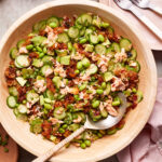 A wooden bowl filled with a salad containing cucumbers, avocado, edamame, and pieces of salmon. The ingredients are mixed with what appears to be sun-dried tomatoes. A serving spoon with a woven handle rests in the bowl. In the background, there are two forks on a pink napkin.