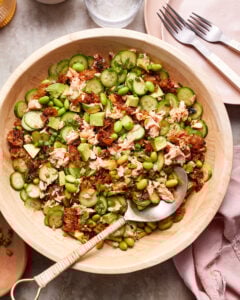 A wooden bowl filled with a salad containing cucumbers, avocado, edamame, and pieces of salmon. The ingredients are mixed with what appears to be sun-dried tomatoes. A serving spoon with a woven handle rests in the bowl. In the background, there are two forks on a pink napkin.