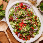 A colorful salad on a white platter featuring slices of beef, cherry tomatoes, sliced cucumbers, red onion, herbs, and sprinkled peanuts. Two glasses with iced drinks are in the background, and a wooden spoon rests nearby.