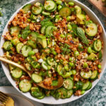 A bowl of fresh salad featuring sliced cucumbers, edamame, avocado chunks, and rice, garnished with chopped green onions and mint leaves. A spoon rests in the bowl, ready to serve. The colorful dish is placed on a green, textured tabletop.