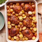 A white baking dish filled with roasted chicken thighs and halved baby potatoes, seasoned and cooked to a golden brown. The dish is on a tiled surface, accompanied by a green plate, a wooden spoon, and a small bowl of salad greens.