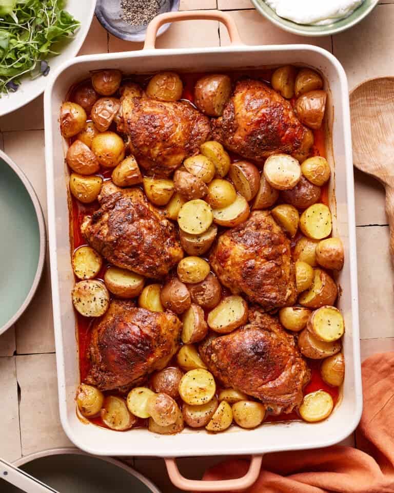 A white baking dish filled with roasted chicken thighs and halved baby potatoes, seasoned and cooked to a golden brown. The dish is on a tiled surface, accompanied by a green plate, a wooden spoon, and a small bowl of salad greens.