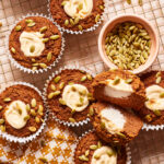 A cooling rack holds six pumpkin muffins, five of them whole and one cut in half to show the creamy filling. The muffins are topped with pumpkin seeds and are placed next to a small bowl filled with additional pumpkin seeds on a patterned napkin.