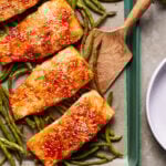 Baked salmon fillets glazed with sauce and sprinkled with sesame seeds, arranged on a baking tray with roasted green beans. A wooden spatula rests nearby.