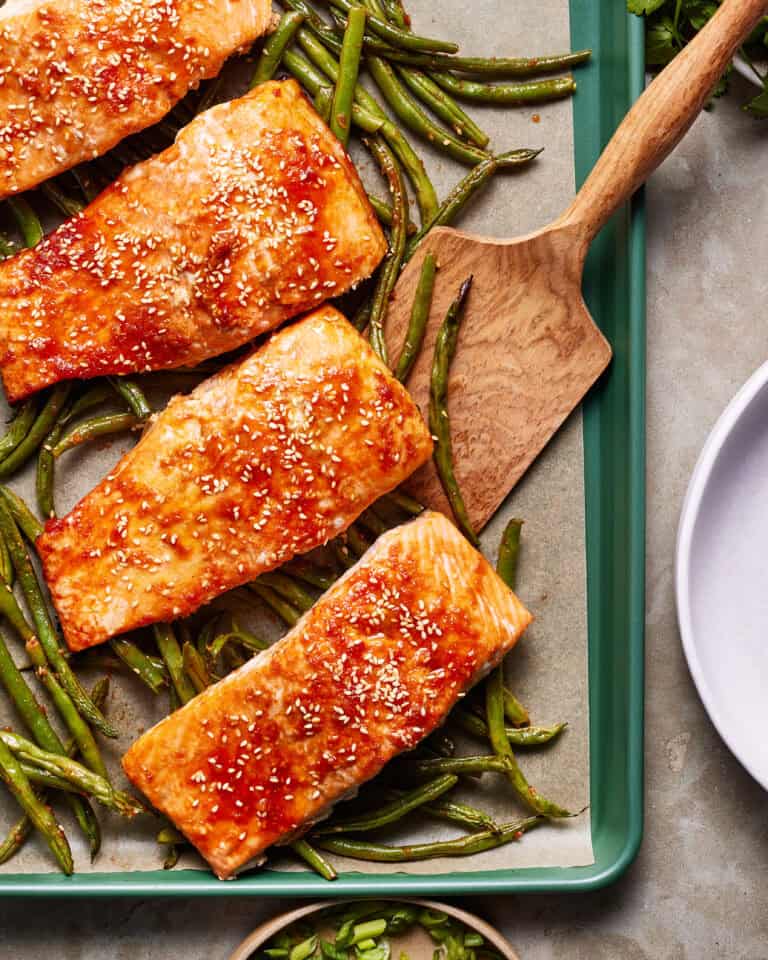 Baked salmon fillets glazed with sauce and sprinkled with sesame seeds, arranged on a baking tray with roasted green beans. A wooden spatula rests nearby.