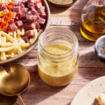 A jar of creamy dressing is positioned on a wooden table, surrounded by a bowl of pasta salad with diced meats, cherry tomatoes, and bell peppers. Nearby are gold utensils, a bottle of olive oil, a small bowl of ground spices, and a piece of bread with grated cheese.