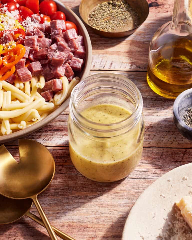 A jar of creamy dressing is positioned on a wooden table, surrounded by a bowl of pasta salad with diced meats, cherry tomatoes, and bell peppers. Nearby are gold utensils, a bottle of olive oil, a small bowl of ground spices, and a piece of bread with grated cheese.