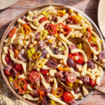 A bowl of pasta salad on a wooden table contains gemelli pasta, cherry tomatoes, pinto beans, black olives, sliced peppers, and crumbled cheese. Two spoons are laid in the bowl, and a woven white and yellow napkin is visible to the left.
