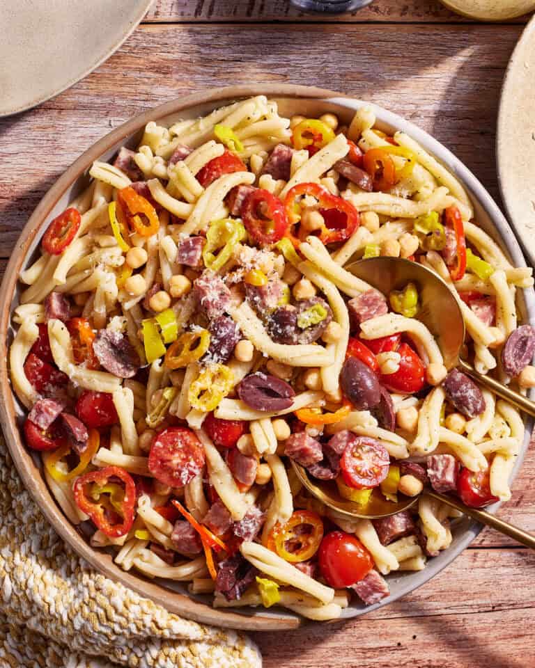A bowl of pasta salad on a wooden table contains gemelli pasta, cherry tomatoes, pinto beans, black olives, sliced peppers, and crumbled cheese. Two spoons are laid in the bowl, and a woven white and yellow napkin is visible to the left.