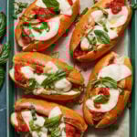 Five caprese hot dogs are arranged on a parchment-lined tray. Each hot dog is topped with slices of mozzarella cheese, tomato sauce, and fresh basil leaves. A few additional basil leaves are scattered around the tray.
