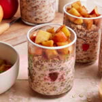 Two glass jars filled with oats and milk, topped with cinnamon-spiced apple chunks. A small red heart decorates each jar. In the background, more jars and a whole red apple are visible on a wooden surface.