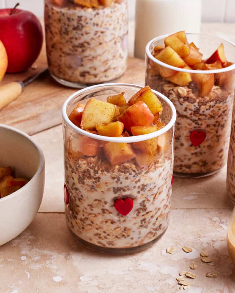 Two glass jars filled with oats and milk, topped with cinnamon-spiced apple chunks. A small red heart decorates each jar. In the background, more jars and a whole red apple are visible on a wooden surface.