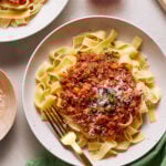 A white plate of pasta topped with a rich red sauce garnished with grated cheese and chopped herbs. A gold fork rests on the plate's edge. A glass of red wine and napkin are nearby.