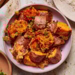 A bowl of crispy roasted sweet potatoes garnished with chopped green onions. The chunks are golden brown with a textured, crispy coating. They sit on a light background, creating an appetizing presentation.