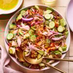 A colorful salad with shredded chicken, sliced cucumbers, red onions, avocado, tomatoes, and crispy bits served on a plate. Two gold serving spoons rest on the salad. A glass of white wine and a pink napkin are nearby.