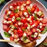 A salad featuring cherry tomatoes, mozzarella pearls shaped like skulls, and fresh basil leaves, served in a white bowl. A gold spoon rests by the salad.