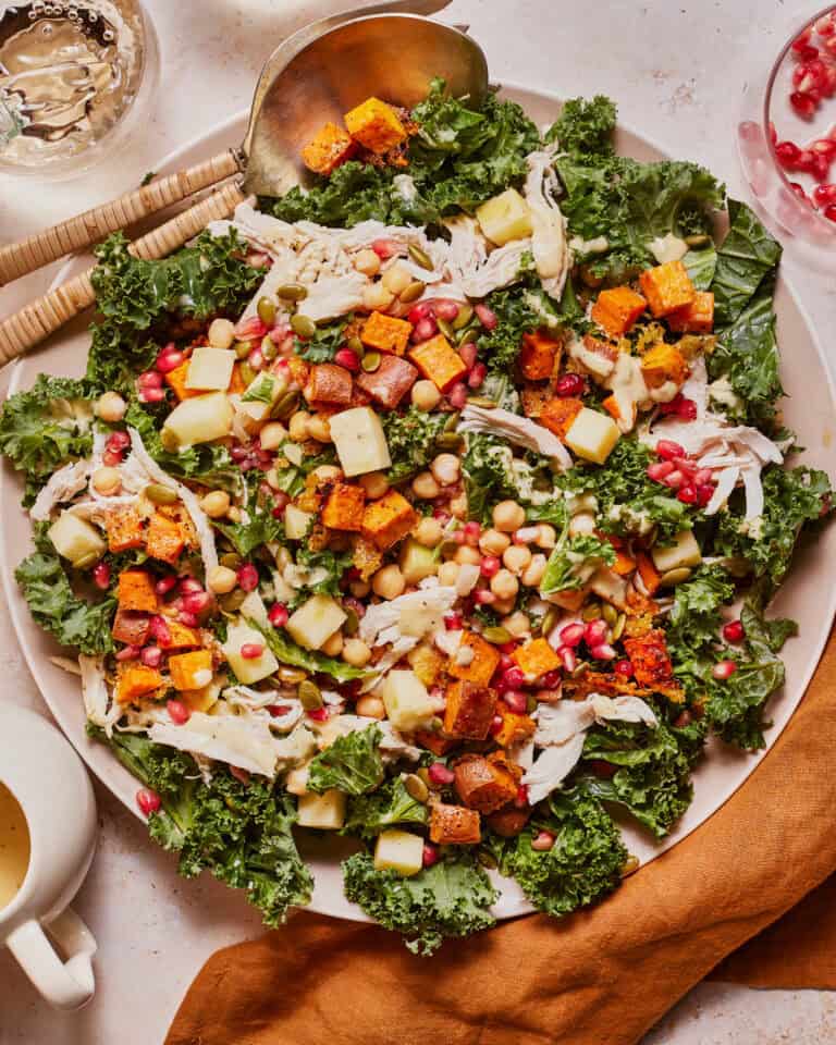 A vibrant salad with shredded chicken, kale, roasted butternut squash, chickpeas, apple cubes, and pomegranate seeds. It is served on a white plate with wooden utensils and a brown napkin beside it.