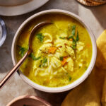 A bowl of chicken orzo soup with pieces of chicken, carrots, and herbs, garnished with fresh parsley. A spoon rests in the bowl. The soup is on a stone surface with a yellow napkin and additional herbs nearby.