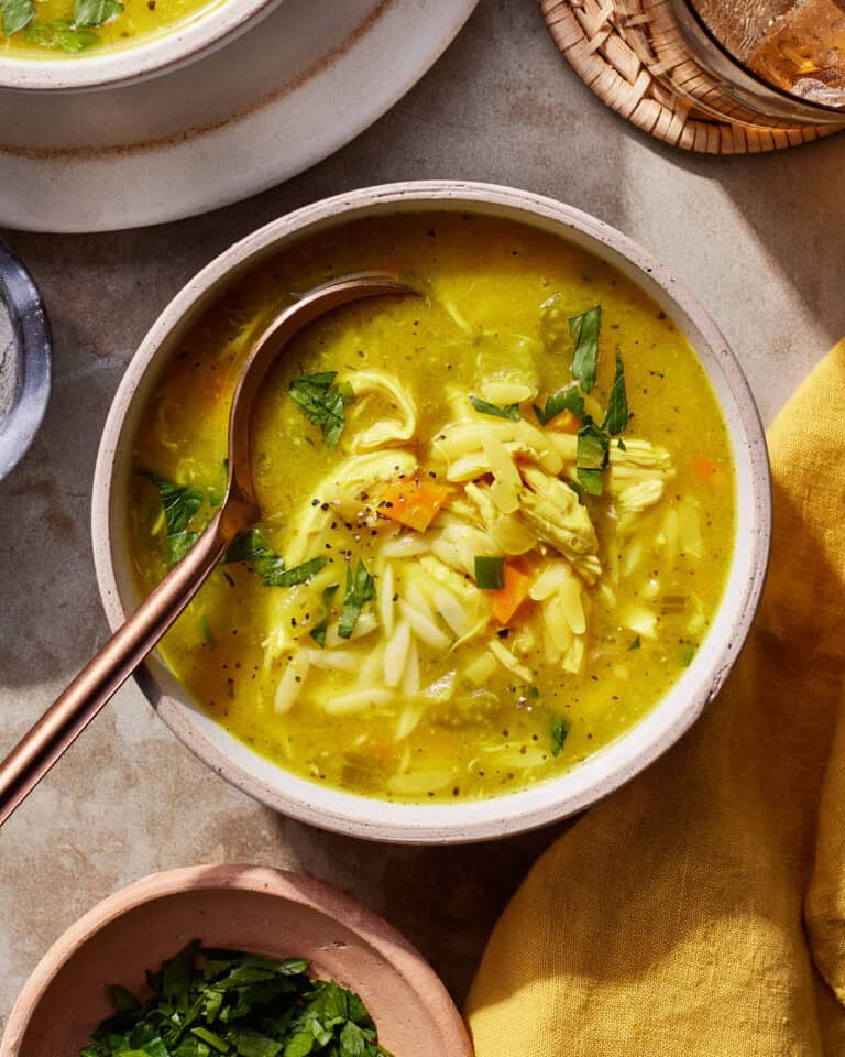 A bowl of chicken orzo soup with pieces of chicken, carrots, and herbs, garnished with fresh parsley. A spoon rests in the bowl. The soup is on a stone surface with a yellow napkin and additional herbs nearby.